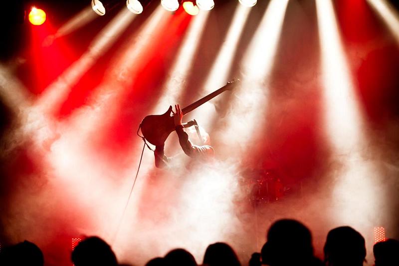 Sylwetka osoby z gitarą na czerwonej scenie, zakryta dymem i ostrymi światłami reflektorów