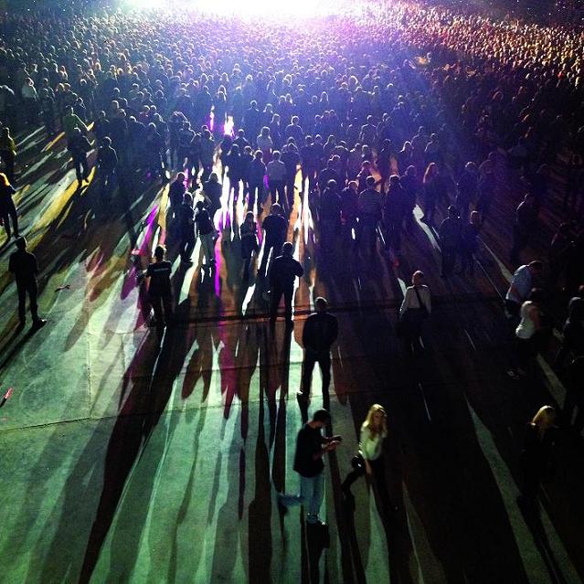 Tłum ludzi stojący przed słupem światła koncertu, za nimi długo rozciągające się cienie