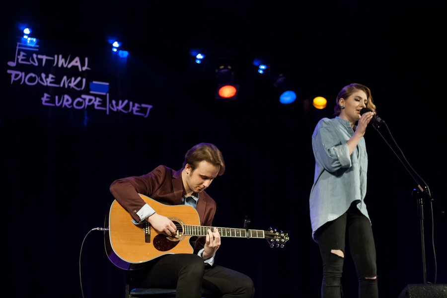 Mężczyzna grający na gitarze, obok niego kobieta w błękitnej bluzce śpiewająca do mikrofonu
