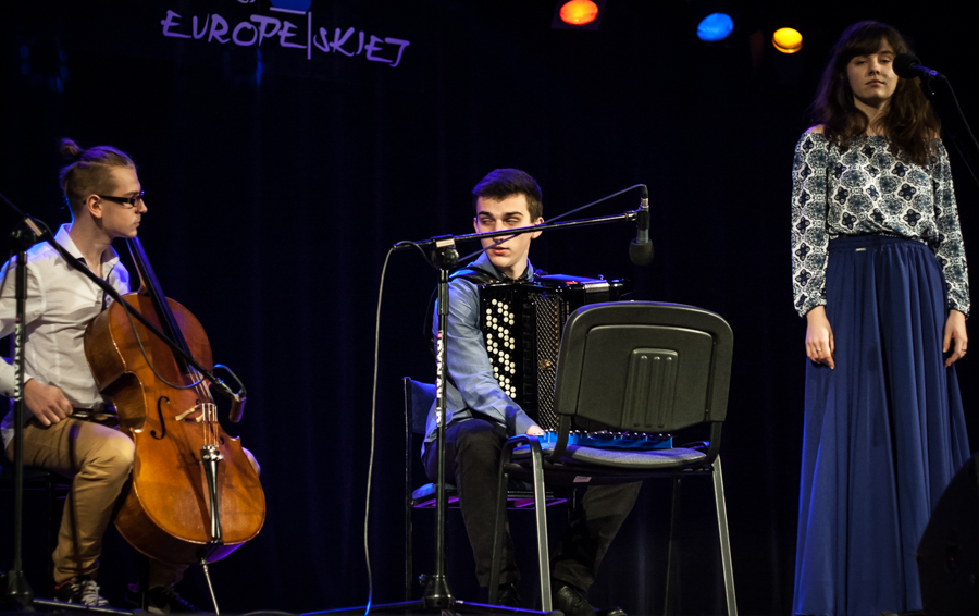 Dwoje mężczyzn grających na instrumentach, obok nich kobieta stojąca przy mikrofonie