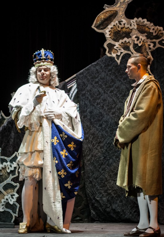 Mężczyzna przebrany za króla w niebiesko-złotych szatach rozmawiający z drugim mężczyzną ubranym w skromny, beżowy płaszcz