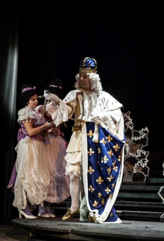 Aktor przebrany za króla w niebiesko-złotych szatach rozmawiający z aktorką w liliowej sukience