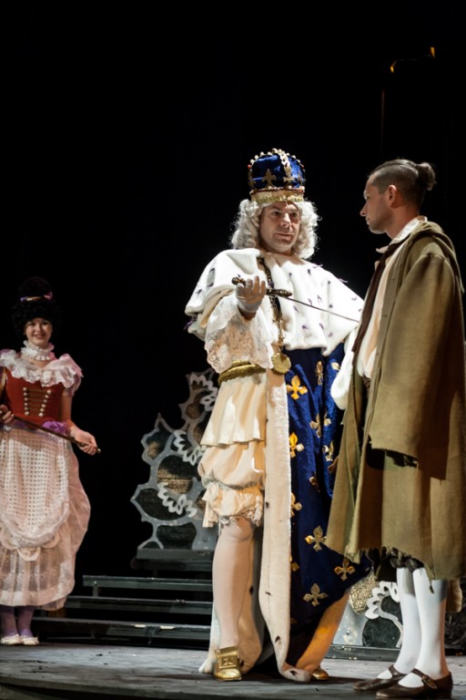 Aktor przebrany za króla w niebiesko-złotych szatach pokazujący szable aktorowi przebranemu w beżowy płaszcz, za nimi stoi aktorka