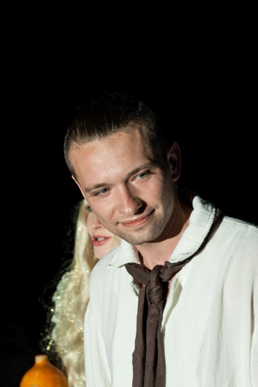 Aktor w białej koszuli i kucyku uśmiecha się, za nim aktorka z długimi blond włosami