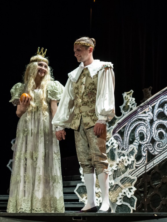 Aktorka z długimi blond włosami trzyma za rękę aktora i w drugiej ręce okrągły przedmiot, na głowie ma koronę, oboje są ubrani w bajeczne, królewskie stroje