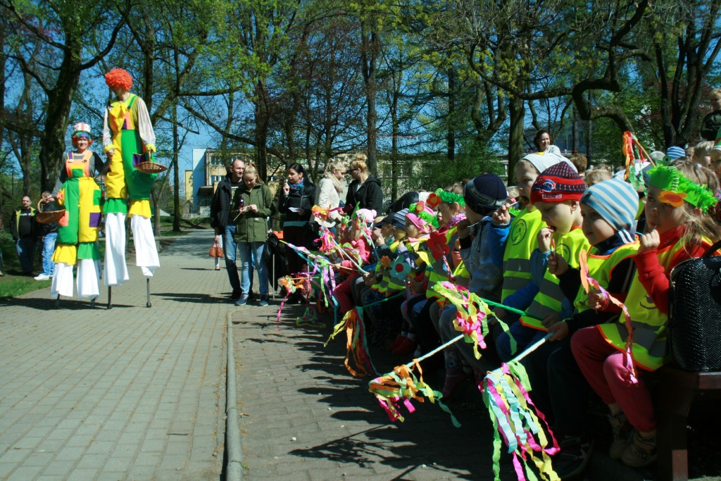 Widownia dzieci w parku trzymające patyki owinięte kolorową bibułą, wszystkie są ubrane w kamizelki odblaskowe, po chodniku idą dwie barwnie ubrane osoby na szczudłach