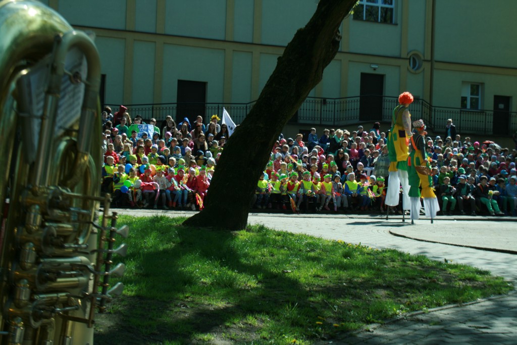Widownia dzieci w parku trzymające patyki owinięte kolorową bibułą, wszystkie są ubrane w kamizelki odblaskowe, po chodniku idą dwie barwnie ubrane osoby na szczudłach, zdjęcie zrobione z oddali, widownie przysłania drzewo
