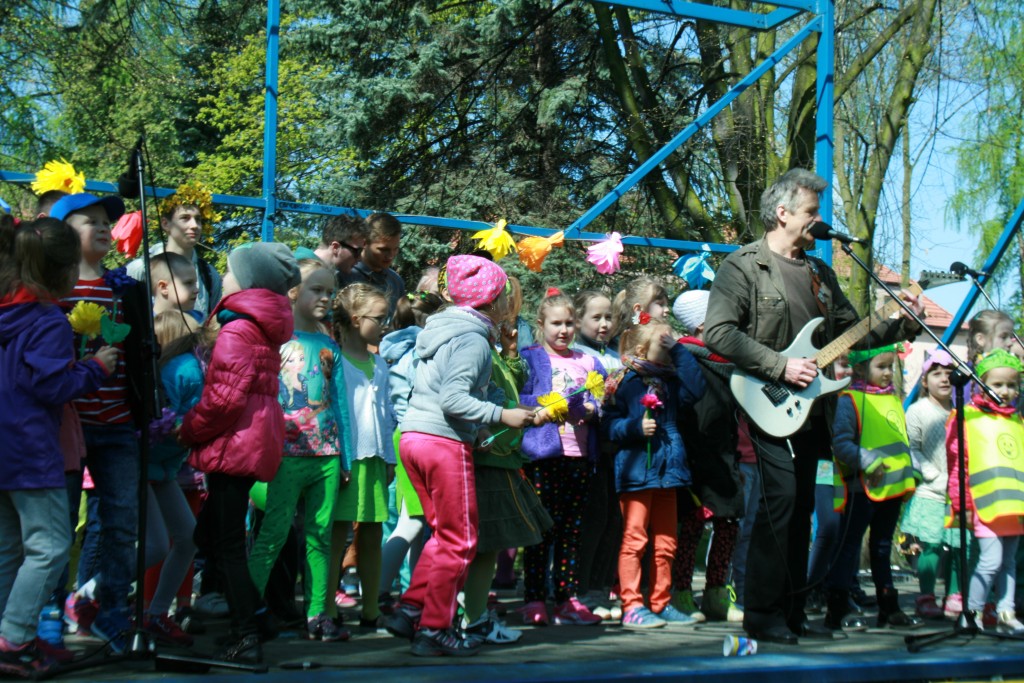 Dzieci stojące na scenie w parku, przed nimi mężczyzna śpiewający do mikrofonu podczas grania na gitarze
