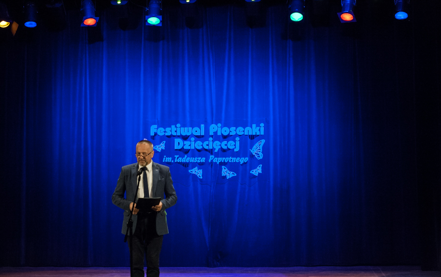 Mężczyzna w garniturze przemawiający na scenie, za nim niebieska kurtyna z napisem 'Festiwal Piosenki Dziecięcej im. Tadeusza Paprotnego'