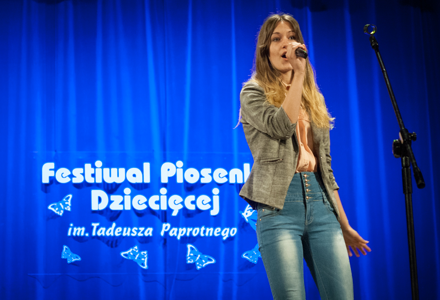 Kobieta śpiewająca do mikrofonu, za nią niebieska kurtyna z napisem 'Festiwal Piosenki Dziecięcej im. Tadeusza Paprotnego'