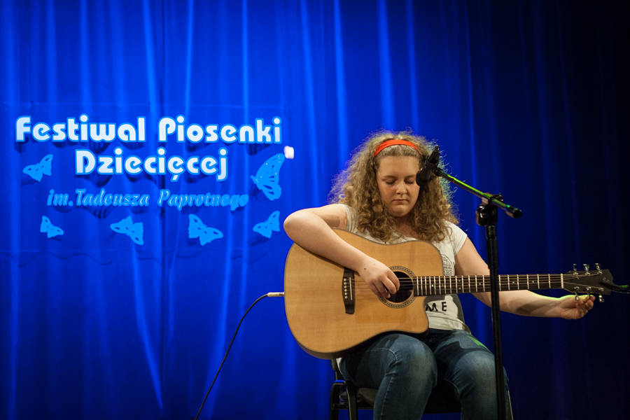Dziewczyna z bujnymi lokami grająca na gitarze, za nią niebieska kurtyna z napisem 'Festiwal Piosenki Dziecięcej im. Tadeusza Paprotnego'