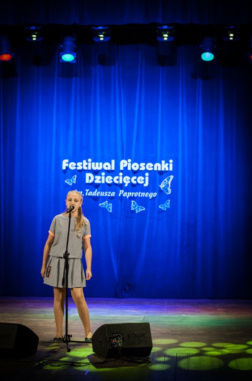 Dziewczyna w szarych ubraniach śpiewająca do mikrofonu, za nią niebieska kurtyna z napisem 'Festiwal Piosenki Dziecięcej im. Tadeusza Paprotnego'