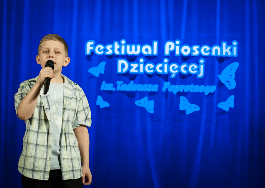 Chłopak w kratkowanej koszuli śpiewający do mikrofonu, za nim niebieska kurtyna z napisem 'Festiwal Piosenki Dziecięcej im. Tadeusza Paprotnego'