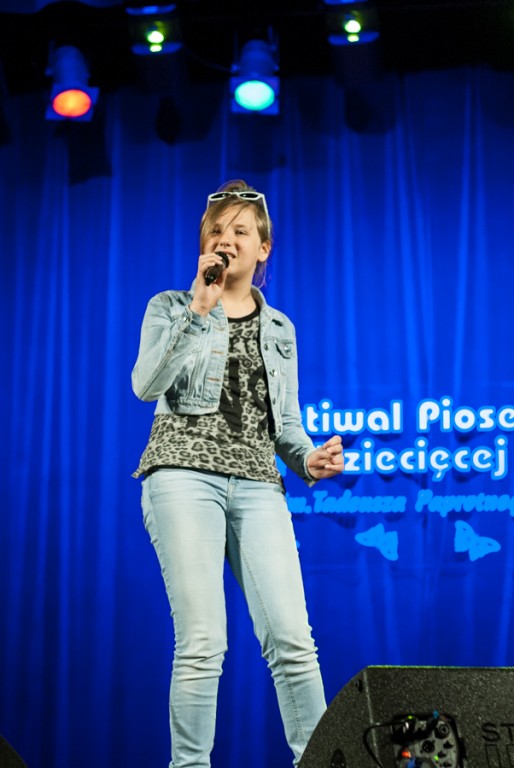 Dziewczyna z założonymi na głowie okularami przeciwsłonecznymi śpiewa do mikrofonu, za nią niebieska kurtyna z napisem 'Festiwal Piosenki Dziecięcej im. Tadeusza Paprotnego'