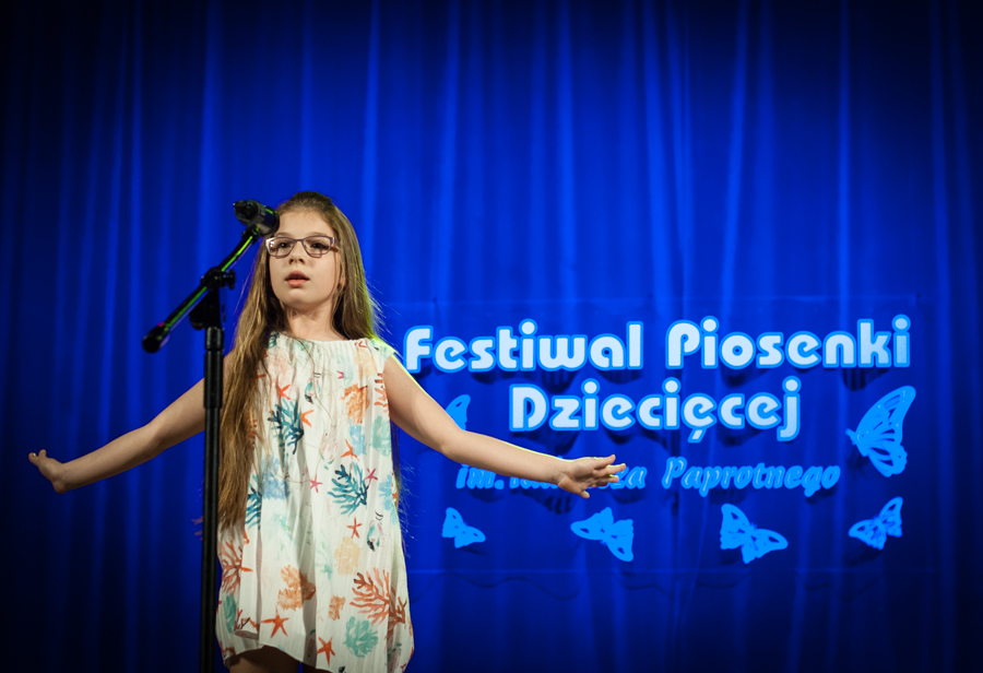Dziewczynka z długimi włosami i okularami śpiewa do mikrofonu, ma rozłożone na boki ręce, za nią niebieska kurtyna z napisem 'Festiwal Piosenki Dziecięcej im. Tadeusza Paprotnego'