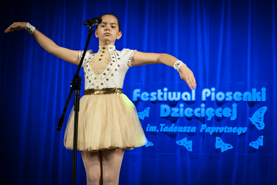 Dziewczynka z tiulowej spódnicy przed mikrofonem wykonuje fale rękami, za nią niebieska kurtyna z napisem 'Festiwal Piosenki Dziecięcej im. Tadeusza Paprotnego'