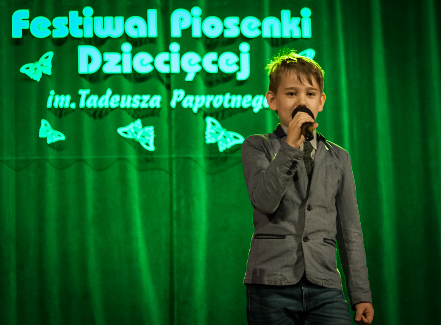 Chłopczyk w garniturze śpiewający do mikrofonu, za nim zielona kurtyna z napisem 'Festiwal Piosenki Dziecięcej im. Tadeusza Paprotnego'