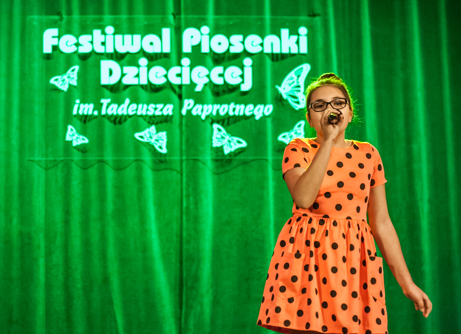 Dziewczyna w sukience w kropki śpiewa do mikrofonu, za nią zielona kurtyna z napisem 'Festiwal Piosenki Dziecięcej im. Tadeusza Paprotnego'