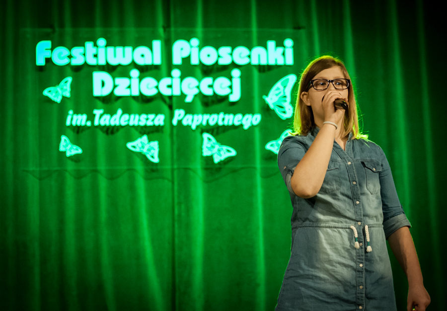 Dziewczyna ubrana w dżins śpiewa do mikrofonu, za nią zielona kurtyna z napisem 'Festiwal Piosenki Dziecięcej im. Tadeusza Paprotnego'