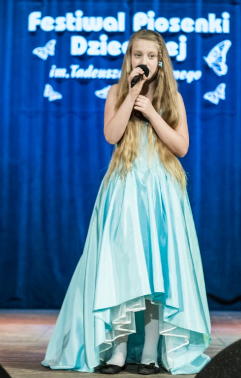 Dziewczynka w błękitnej sukience śpiewająca do mikrofonu, za nią niebieska kurtyna z napisem 'Festiwal Piosenki Dziecięcej im. Tadeusza Paprotnego'