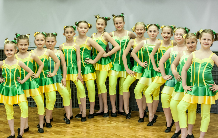 Czternaście dziewczynek ubranych w zielono-żółte sukienki, ustawione obok siebie z rękami na biodrach