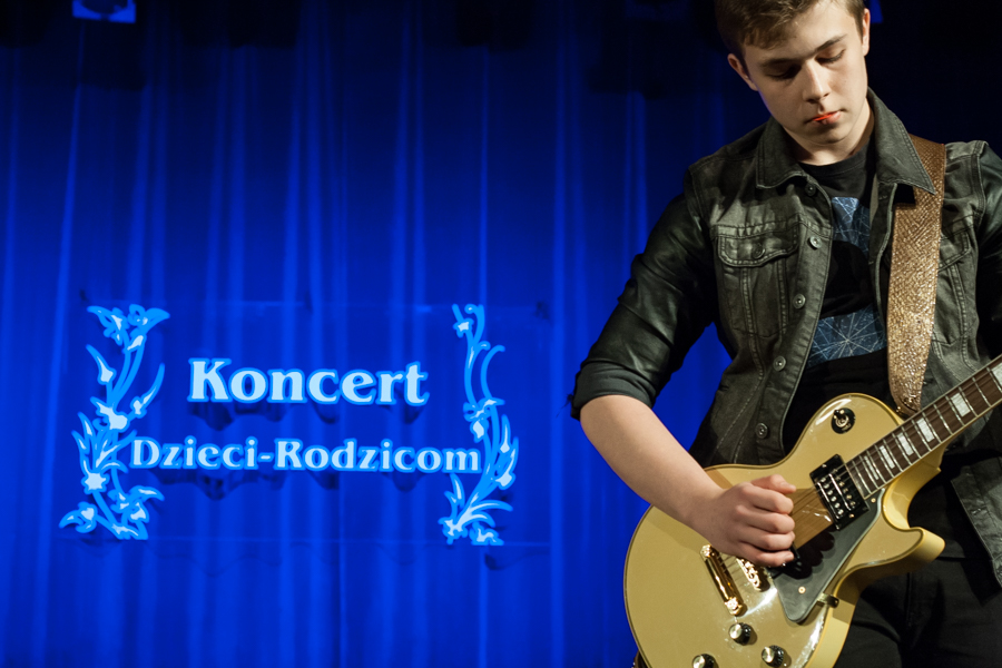 Chłopak w skórzanej kurtce grający na gitarze, za nim niebieska kurtyna z napisem 'Koncert Dzieci-Rodzicom'