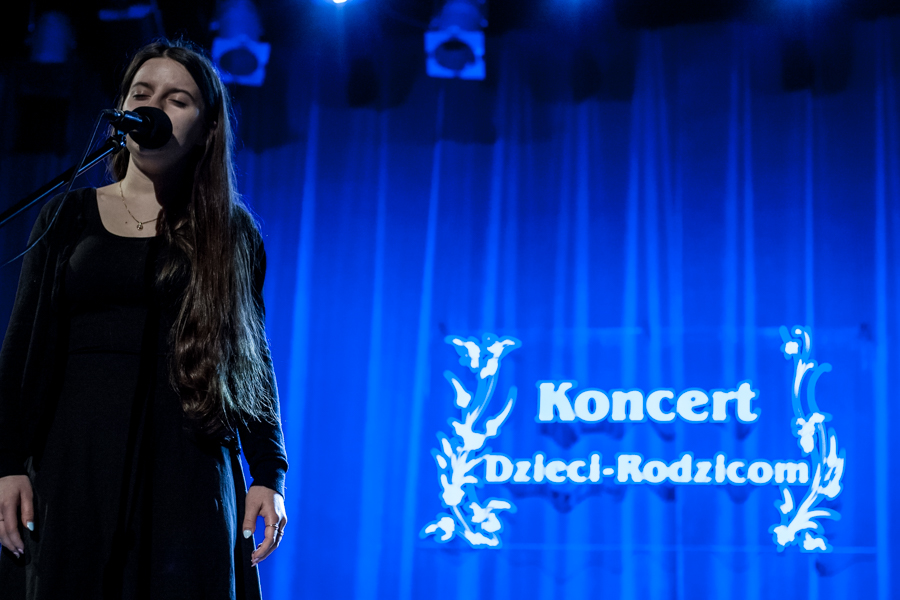 Dziewczyna w czarnej sukience śpiewa do mikrofonu, za nią niebieska kurtyna z napisem 'Koncert Dzieci-Rodzicom'
