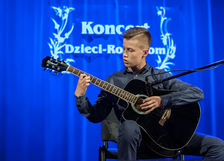 Chłopak grający na czarnej gitarze, za nim niebieska kurtyna z napisem 'Koncert Dzieci-Rodzicom'