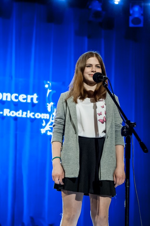 Dziewczyna z jasno-niebieskim swetrze przed mikrofonem, za nią niebieska kurtyna z napisem 'Koncert Dzieci-Rodzicom'