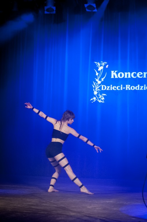 Kobieta z czarnymi paskami na rękach i nogach tańcząca na scenie, za nią niebieska kurtyna z napisem 'Koncert Dzieci-Rodzicom'