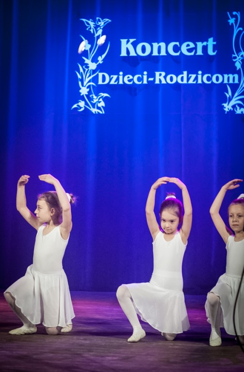 Trzy dziewczynki ubrane w białe sukienki tańczą na scenie, za nimi niebieska kurtyna z napisem 'Koncert Dzieci-Rodzicom'