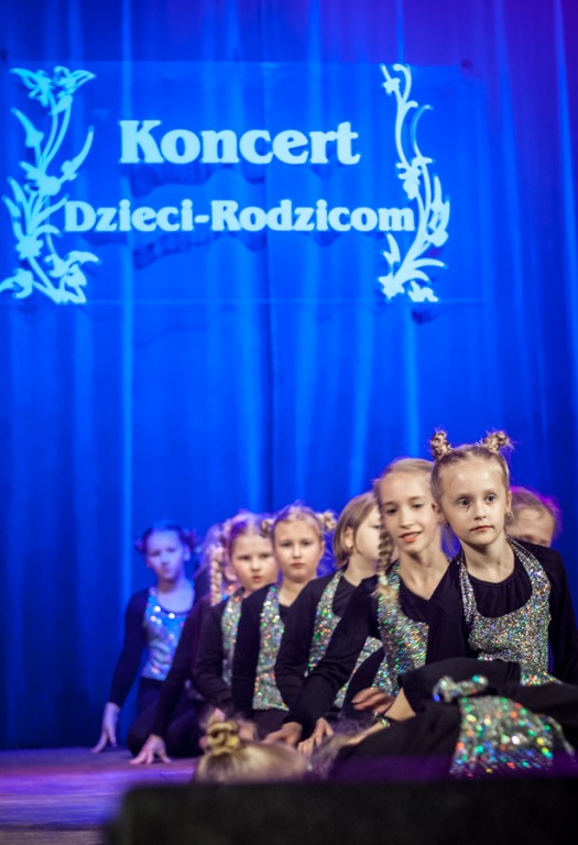 Dziewczynki ubrane w czarno-brokatowe sukienki siedzą gęsiego na scenie, za nimi niebieska kurtyna z napisem 'Koncert Dzieci-Rodzicom'