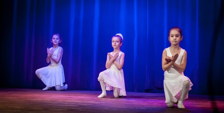 Trzy dziewczynki ubrane w białe sukienki występują na scenie, za nimi niebieska kurtyna