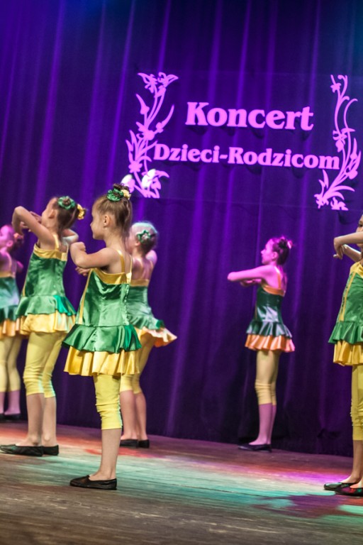 Dziewczynki ubrane w zielono-żółte sukienki występują na scenie, za nimi fioletowa kurtyna z napisem 'Koncert Dzieci-Rodzicom'
