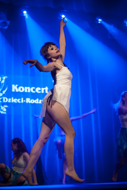 Kobieta ubrana w biel tańczy na scenie, za nią inni występujący na tle niebieskiej kurtyny z napisem 'Koncert Dzieci-Rodzicom'
