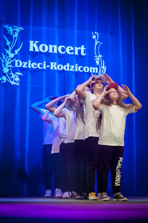 Grupa dziewczynek z czapkami z daszkiem występują na scenie, za nimi niebieska kurtyna z napisem 'Koncert Dzieci-Rodzicom'