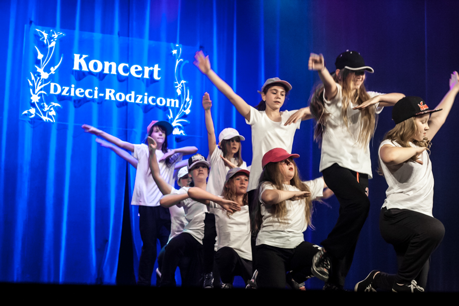 Dziewczynki ubrane w czapki z daszkiem tańczą na scenie, za nimi niebieska kurtyna z napisem 'Koncert Dzieci-Rodzicom'