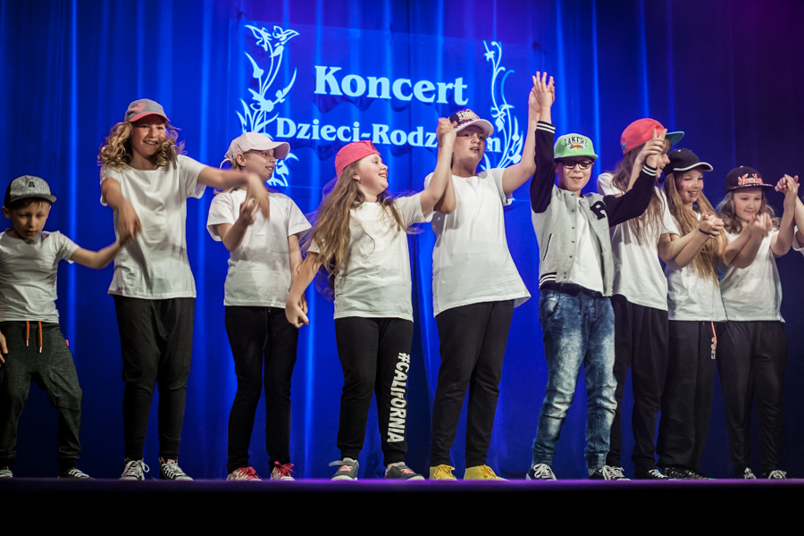 Zdjęcie grupowe tancerzy w czapkach z daszkiem, za nimi niebieska kurtyna z napisem 'Koncert Dzieci-Rodzicom'