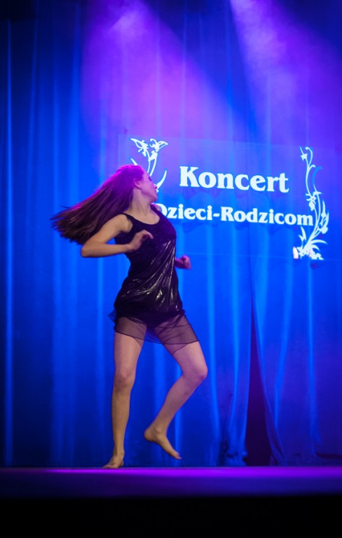 Dziewczyna ubrana w czerń tańczy na scenie, za nią niebieska kurtyna z napisem 'Koncert Dzieci-Rodzicom'