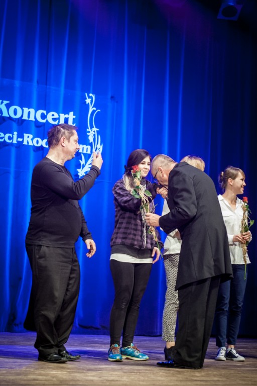 Elegancko ubrany mężczyzna rozdaje nagrody osobom na scenie, całuje dłoń jednej kobiecie, za nimi niebieska kurtyna z napisem 'Koncert Dzieci-Rodzicom'