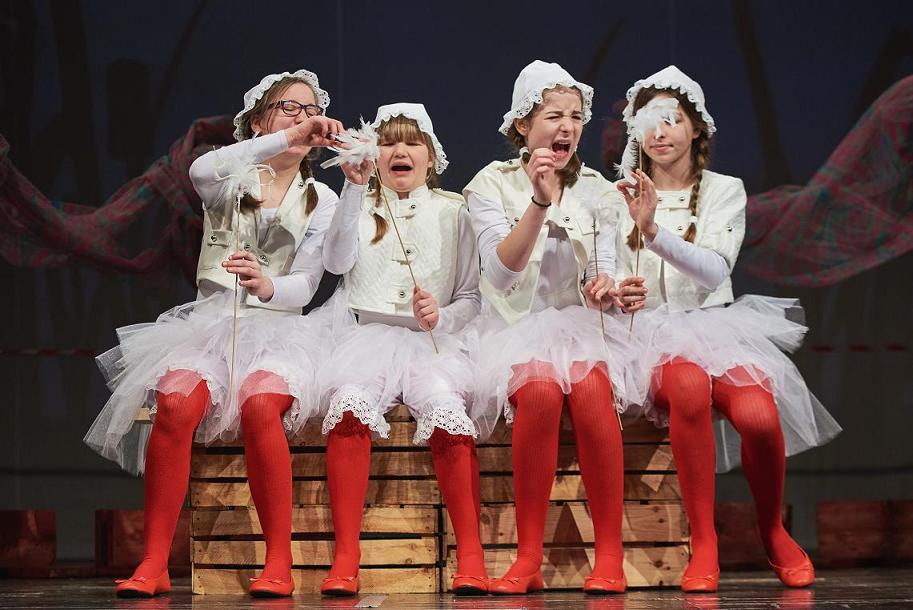 Cztery dziewczyny w warkoczach ubrane w białe sukienki i czapki, z czerwonymi rajstopami siedzą obok siebie na drewnianym pudle