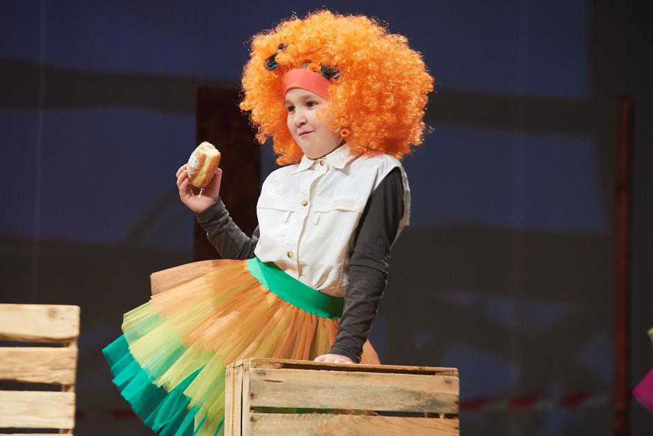Dziewczynka ubrana w kolorową spódnicę i rudą, bujną, kręconą perukę, opierająca się o drewnianą skrzynkę i jedząca pączka
