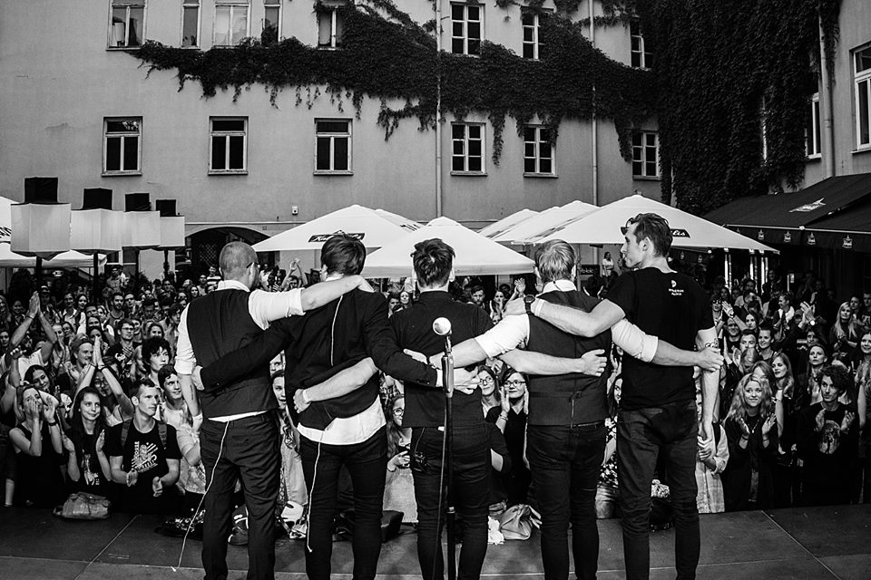 Czarno-białe zdjęcie obejmujących się piątki mężczyzn na scenie przed widownią