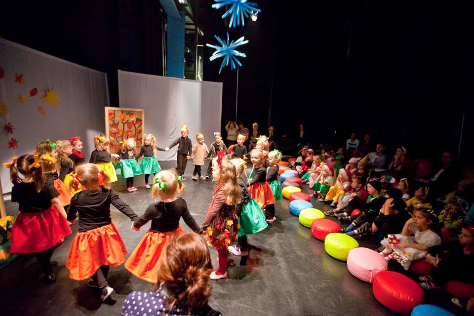 Kolorowo poubierane dzieci na scenie trzymają się za ręce i tańczą w kółku, widownia patrzy na nie z zaciekawieniem