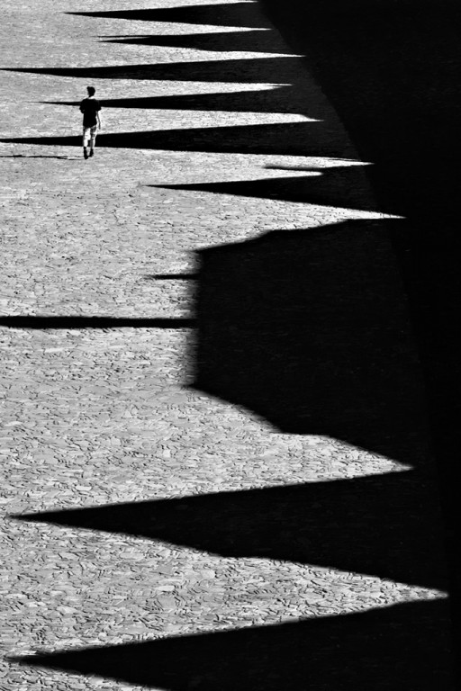 Czarno-białe zdjęcie osoby idącej po chodniku, na nim długie słupy cieni budynków
