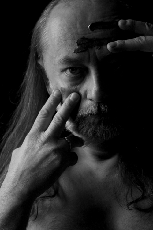 Czarno-białe zdjęcie brodatego mężczyzny z długimi włosami malujący palcami farbą po swojej twarzy