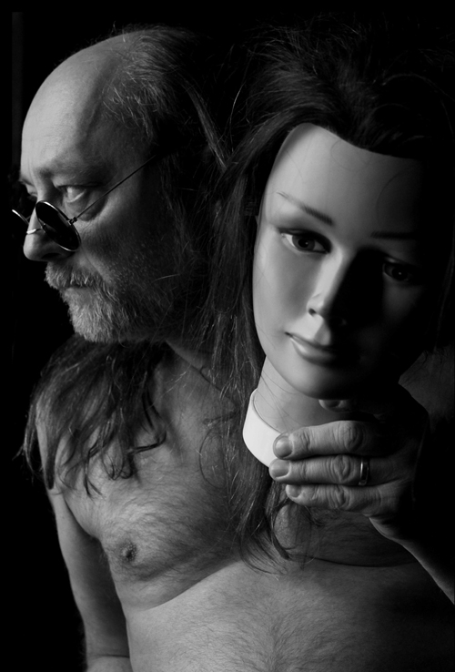 Czarno-białe zdjęcie brodatego mężczyzny w okularach z długimi włosami, trzymającego w lewej dłoni głowę manekina kobiety