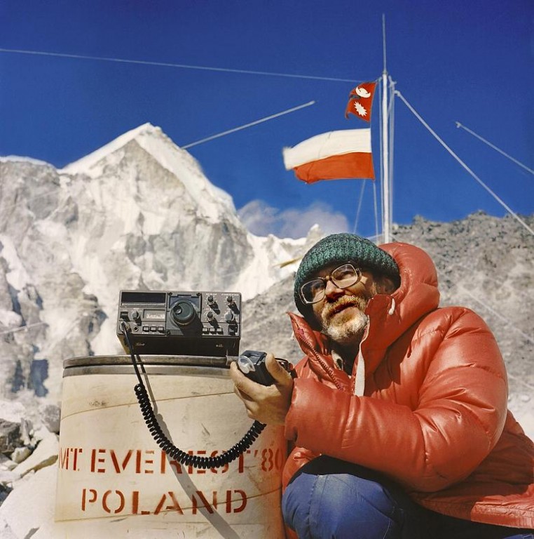Brodaty mężczyzna w czerwonej kurtce trzymający urządzenie, za nim śnieżne góry i polska flaga na słupie