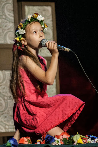 Dziewczynka w wianku i różowej sukience śpiewa do mikrofonu, pod nią porozrzucane sztuczne kwiaty