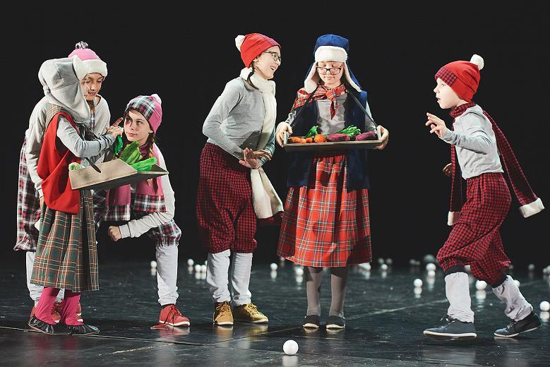 Zimowo poubierane dzieci na scenie grają, rozmawiają i trzymają tace z jedzenie